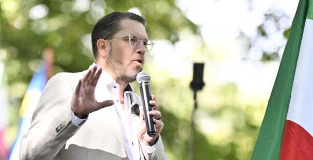 Jimmie Åkesson talar i mikrofon framför en iransk flagga i Humlegården