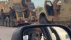 Flera sandfärgade pansarfordon på transporttrailrar medan filmaren syns i en backspegel.