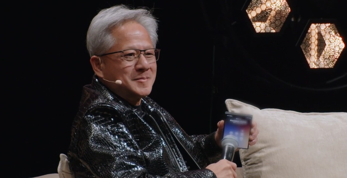 Nvidias vd Jensen Huang sitter på scen i skinnjacka med mikrofon och mobiltelefon i händerna.