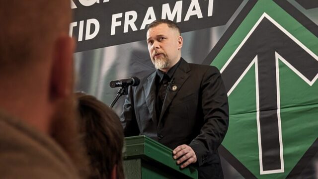 Fredrik Vejdeland talar vid en talarstol med Nordiska motståndsrörelsens gröna pilsymbol bakom sig.
