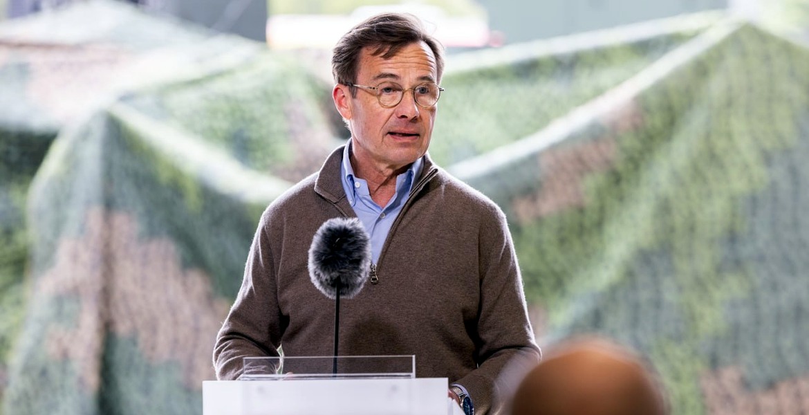 Ulf Kristersson talar vid en talarstol med mikrofon, i brun tröja och glasögon.
