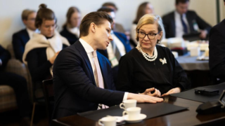 Två personer i samtal vid ett bord under Samlingspartiets riksdagsgrupps möte, med andra deltagare i bakgrunden.