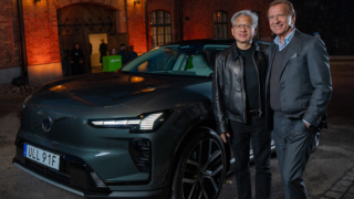 Två män står bredvid en mörk Volvobil framför en varmt belyst tegelfasad.