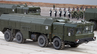 Ett grönmålat Iskander-M-avfyrningsfordon med hela fordonet synligt på en betongyta och militär personal i bakgrunden.