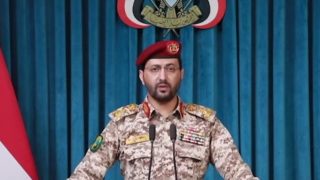Huthiernas militära talesperson Yahya Saree i uniform och röd basker framför mikrofoner och blå draperier.